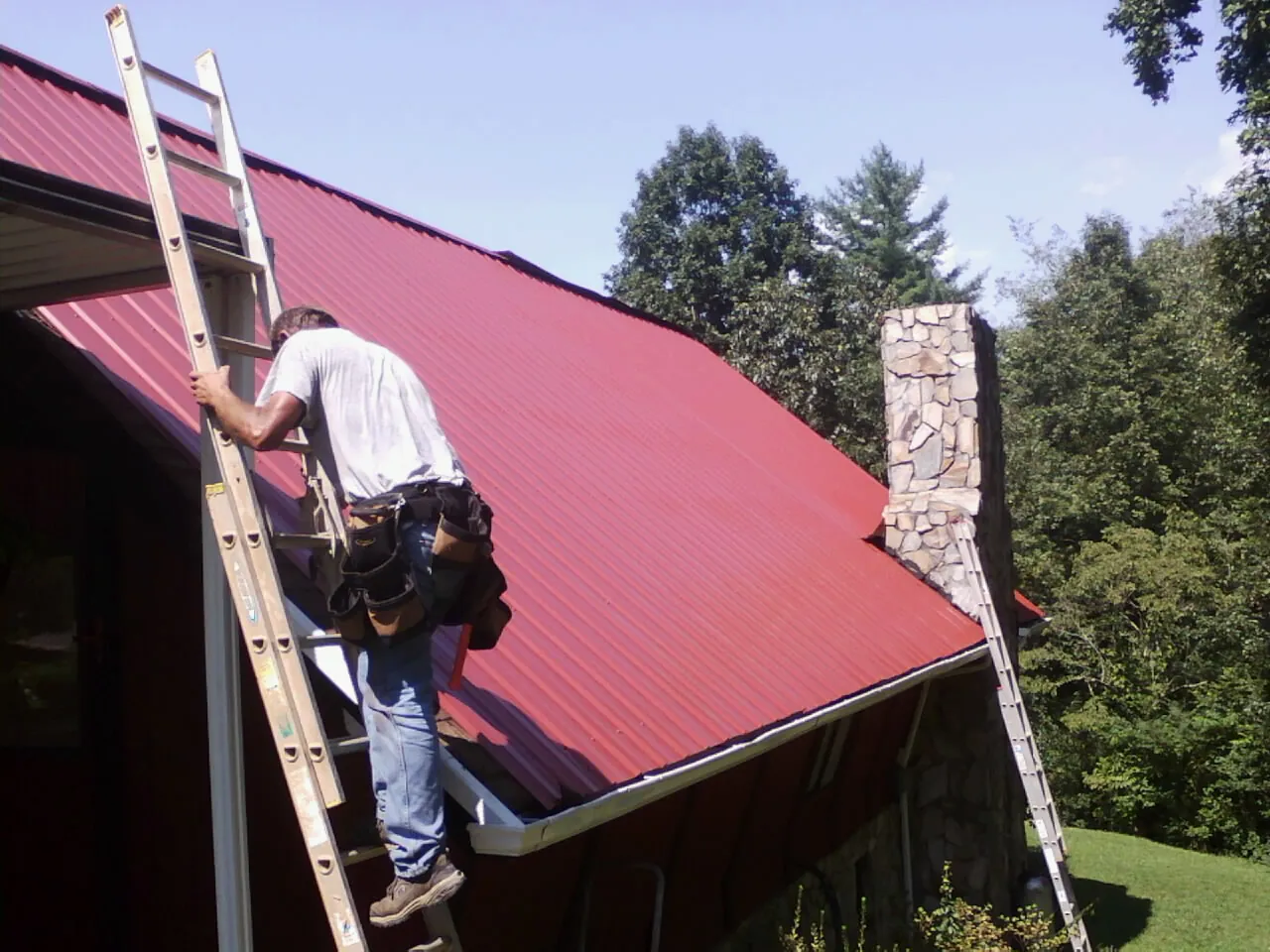 Roofer on a ladder beside a red metal roof and stone chimney — Soffit & Fascia Repair by Roofing Brothers in Weskan